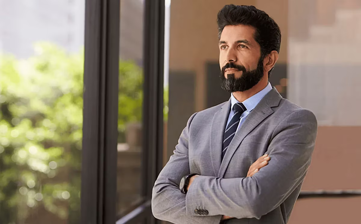 man with beard and grey suit arms crossed smoldering